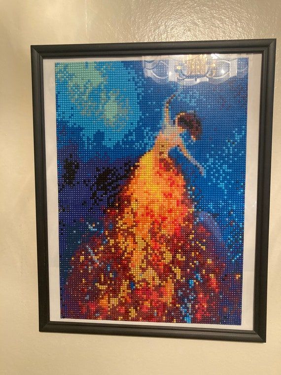 Inramad diamond painting tavla föreställande en kvinna i guldklänning mot en blå bakgrund.